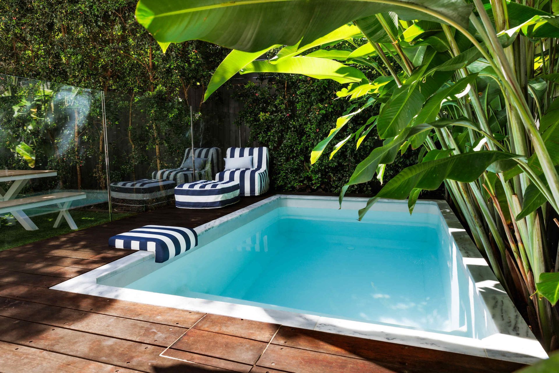 Small pool surrounded by greenery and wooden decking. Striped chairs and ottoman are nearby. Plungie Pools