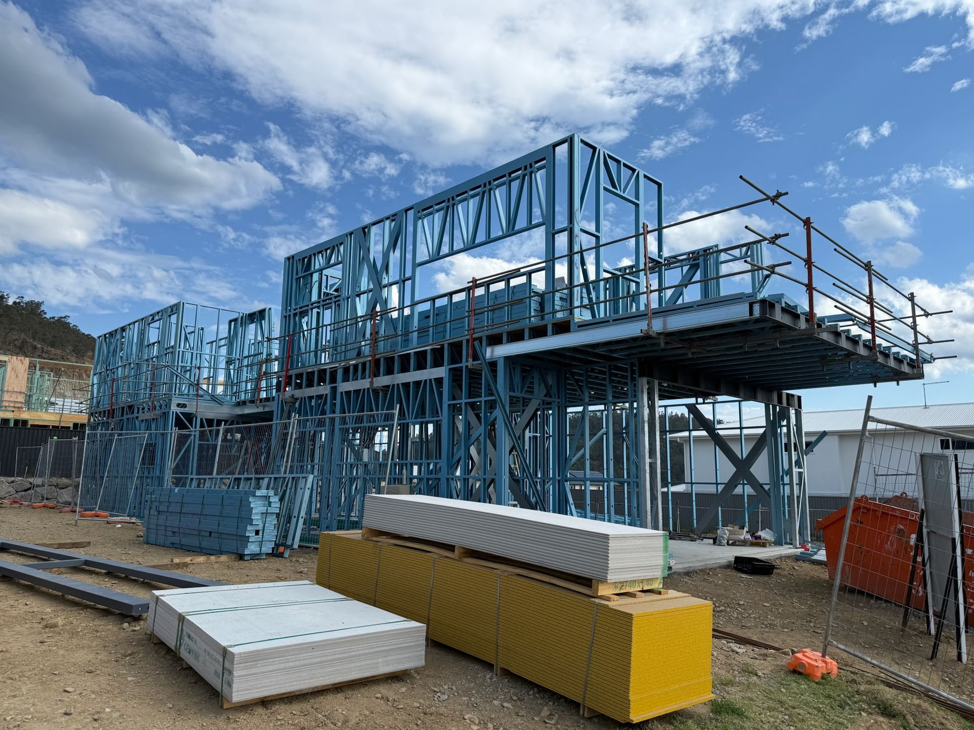 Construction site with blue metal TRUECORE® Steel framing for a building. Materials stacked nearby, against a partly cloudy sky.