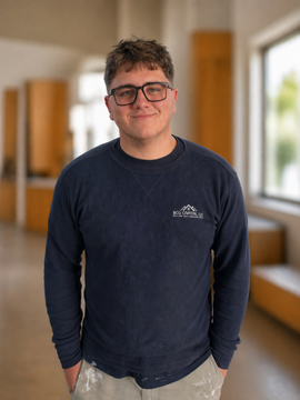 Person standing indoors in a navy shirt and jeans, smiling in a bright hallway.
