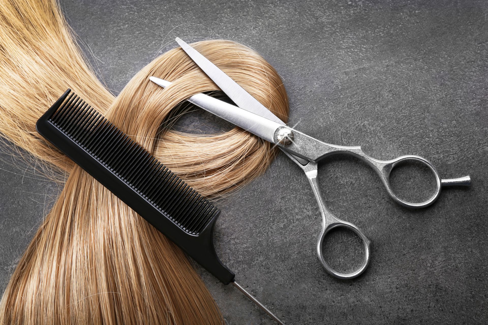 Friseurutensilien: blondes Haar, schwarzer Kamm, Schere und Bindfaden auf grauem Hintergrund