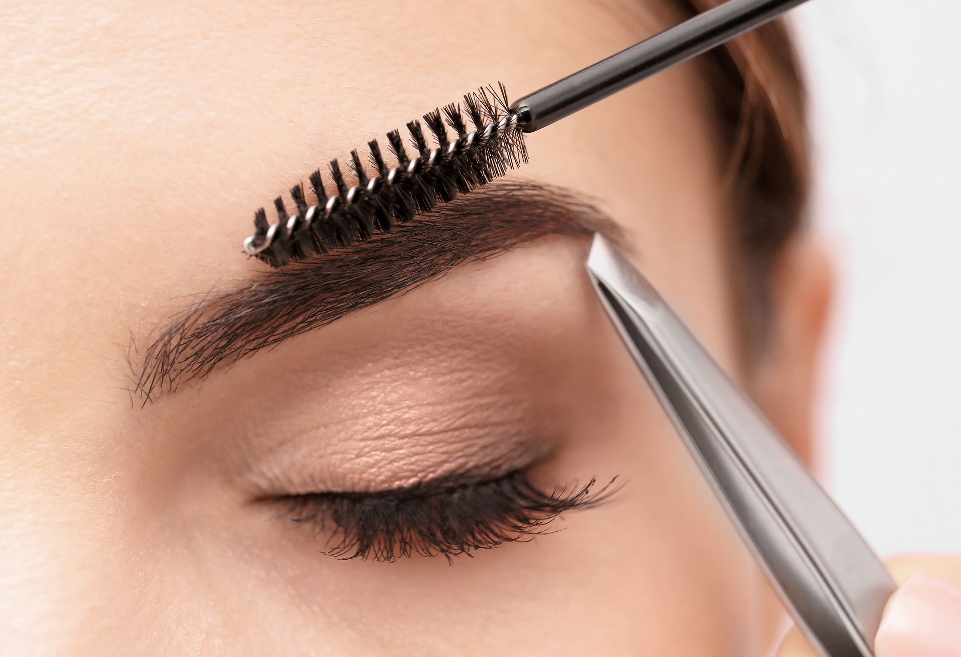 Augenbrauen werden mit einem Wimpernbürstchen und einer Pinzette in der Nähe eines geschlossenen Auges gestylt