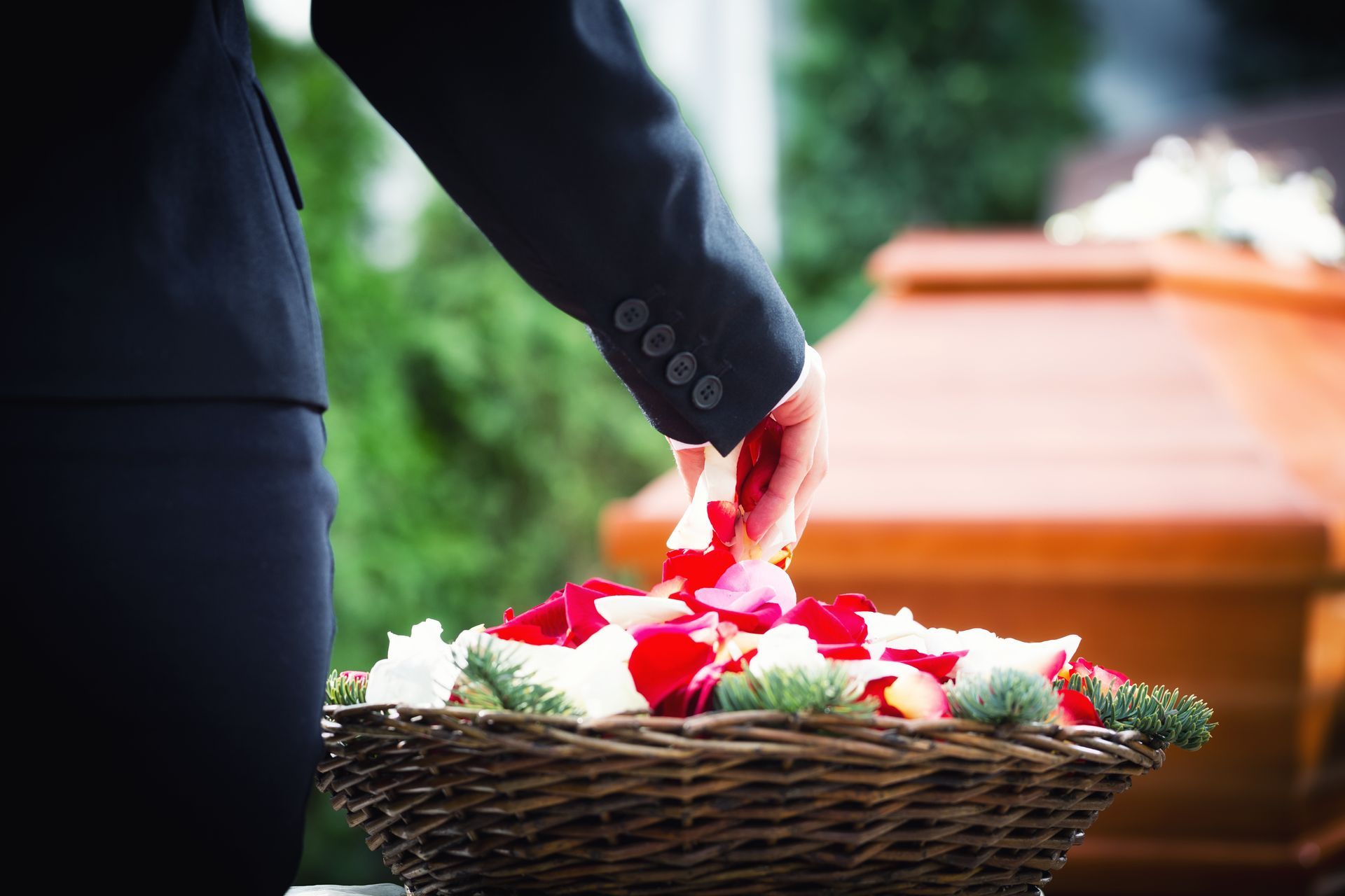 Una persona in abito scuro allunga la mano verso un cesto di vimini pieno di petali di rosa, con una bara di legno sullo sfondo.