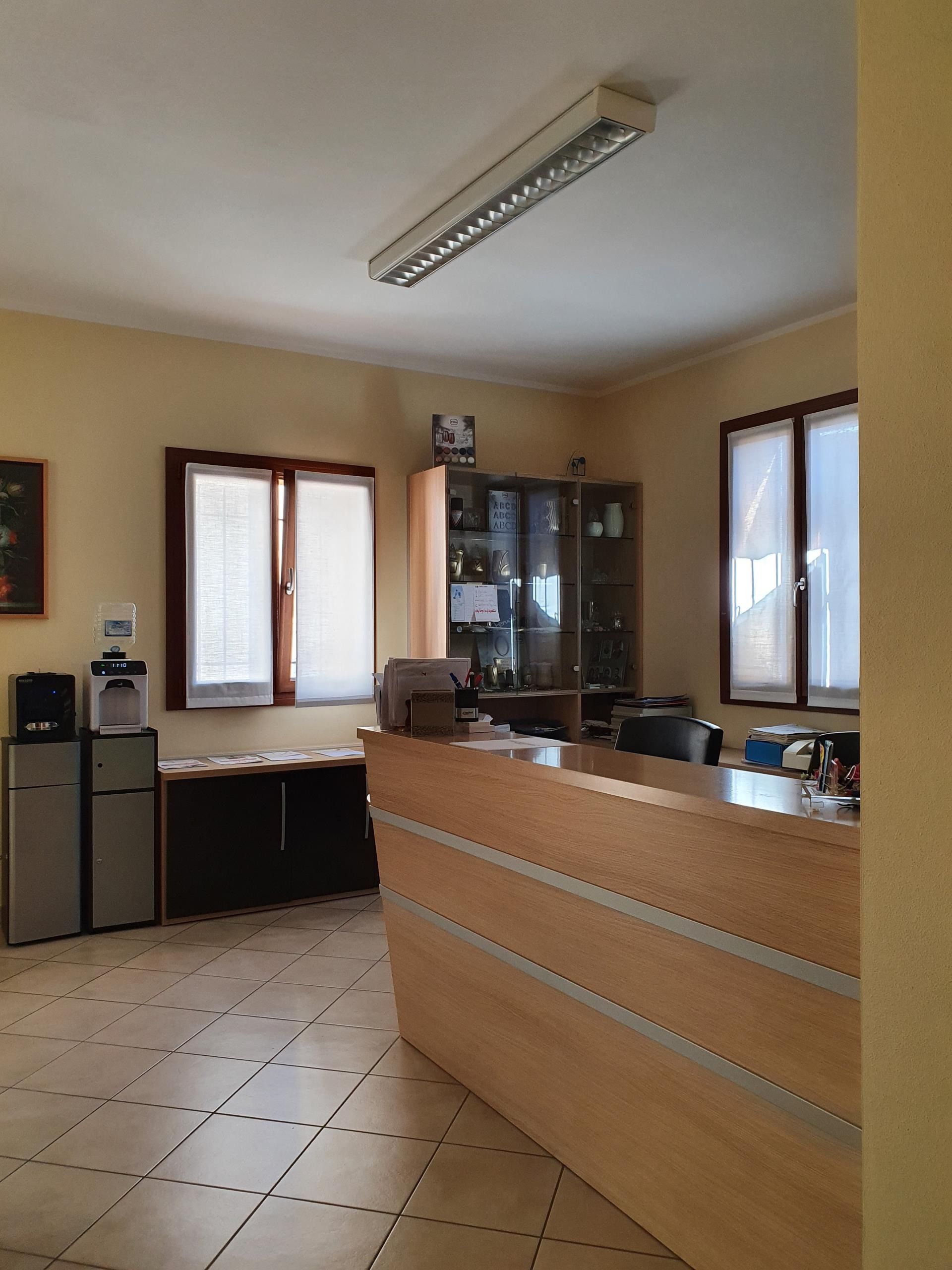 Una zona reception con una scrivania in legno in primo piano, pavimento piastrellato, una vetrina e finestre con tende bianche.