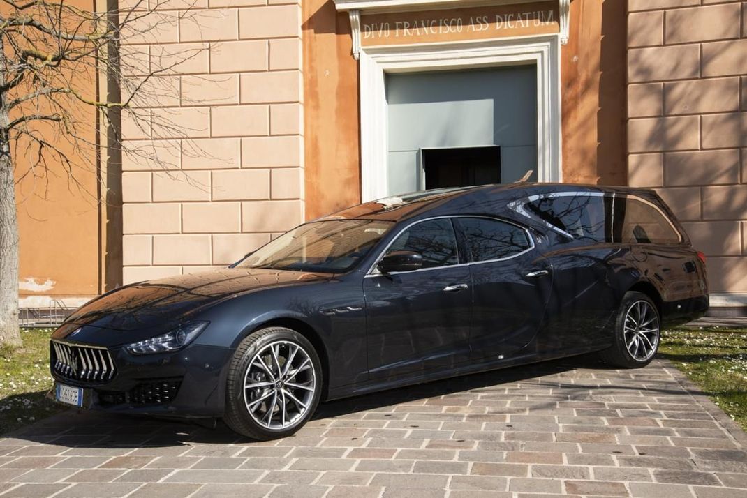 Un carro funebre Maserati di colore scuro parcheggiato davanti a un edificio in mattoni color beige su un patio in pietra.