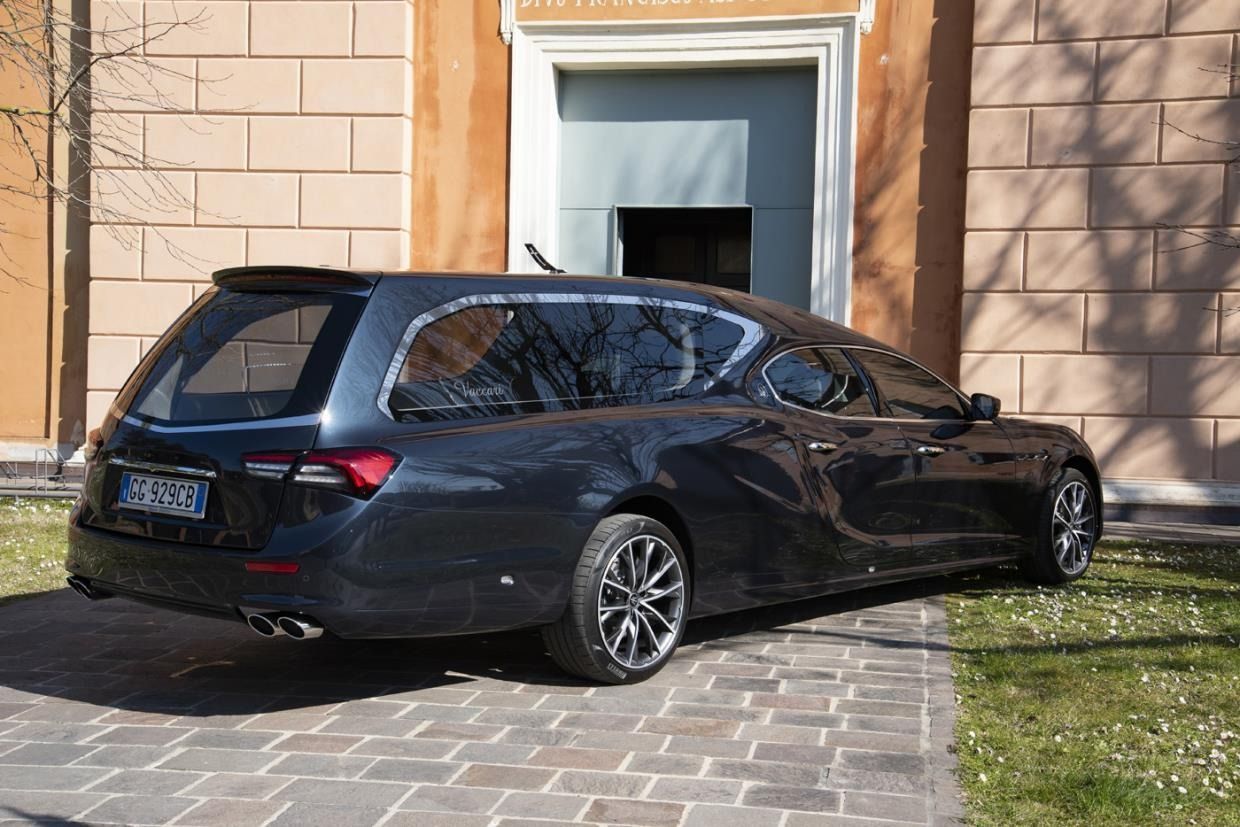 Un carro funebre grigio scuro parcheggiato su un vialetto di pietra di fronte a un edificio in mattoni.