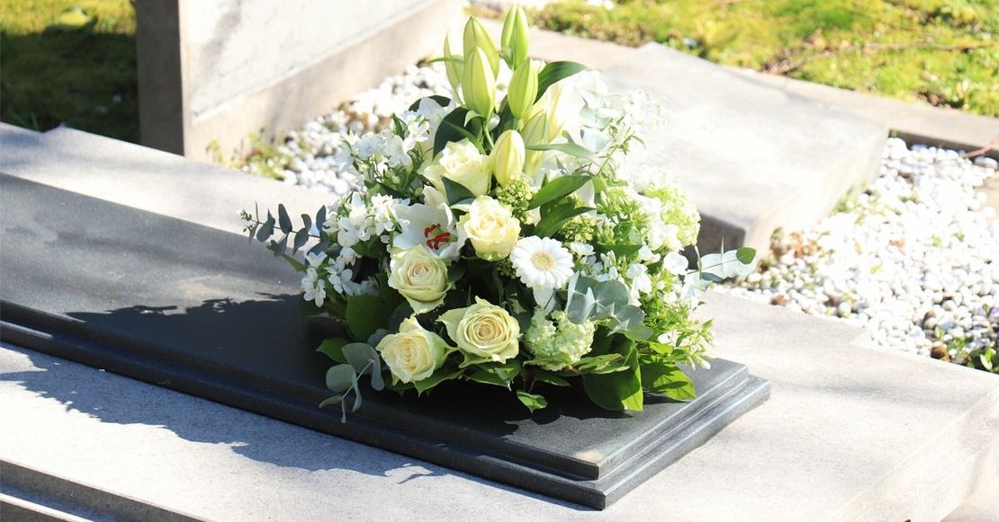 Una composizione floreale di rose bianche, gigli e verde poggiata su una lapide commemorativa in pietra grigia in un cimitero.