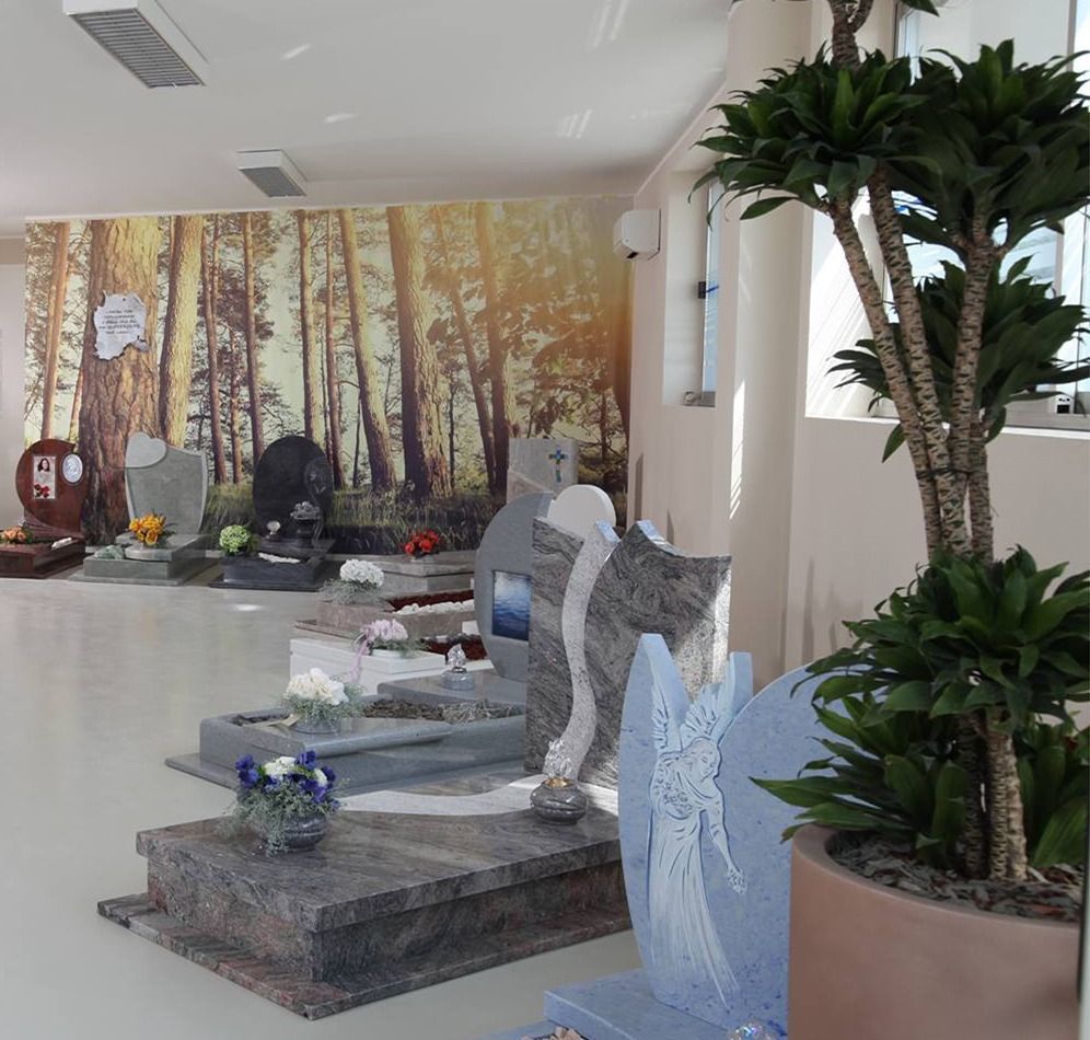 Uno showroom che espone diverse lapidi e monumenti in pietra di fronte a un murale raffigurante una foresta, con una grande pianta in vaso nelle vicinanze.