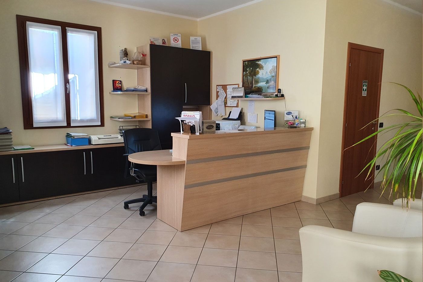 Una zona reception con scrivania in legno chiaro, armadio scuro, sedia da ufficio, finestra e pavimento in piastrelle color crema.