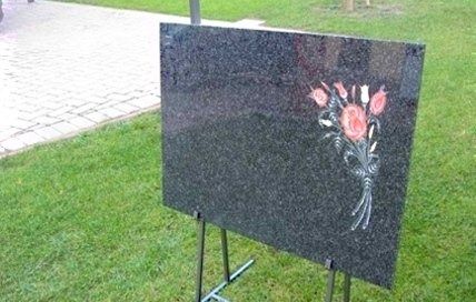 Una lapide in granito nero lucido con un mazzo di fiori inciso, posta su un piedistallo metallico in un'area erbosa.