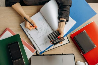 Hands writing on paperwork with a calculator, surrounded by colorful notebooks on a desk.