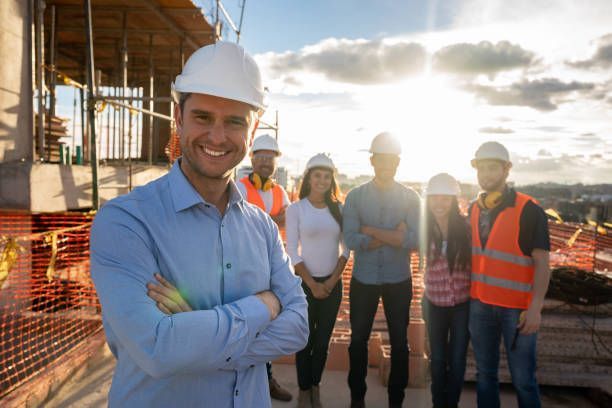 Construction workers with hard hats pose at a sunny construction site, smiling.