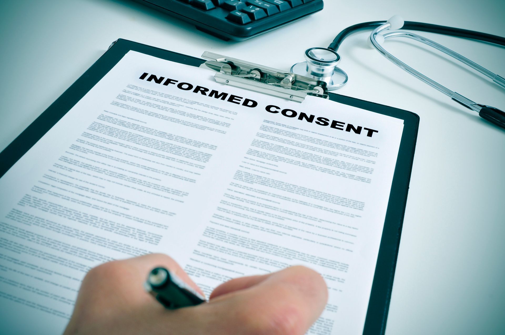 Hand writing on an informed consent form on a clipboard with a stethoscope and keyboard in the background.