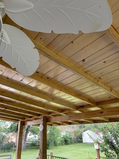 Wooden pergola ceiling with white leaf-shaped fan blades, overlooking a backyard with grass and trees. Wooden pergola ceiling with white leaf-shaped fan blades, overlooking a backyard with grass and trees.