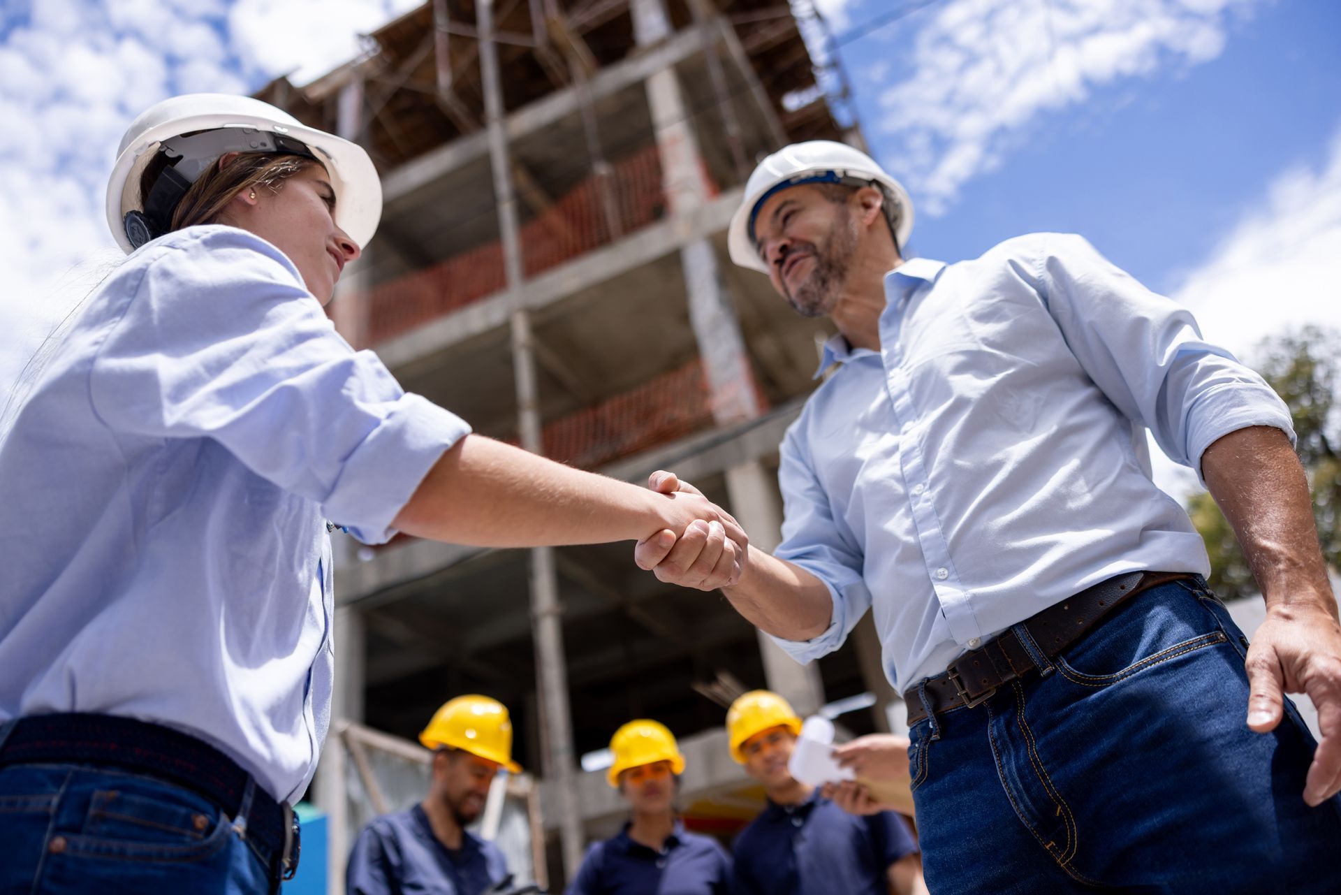 Two construction workers in hard hats shake hands in front of a building under construction.