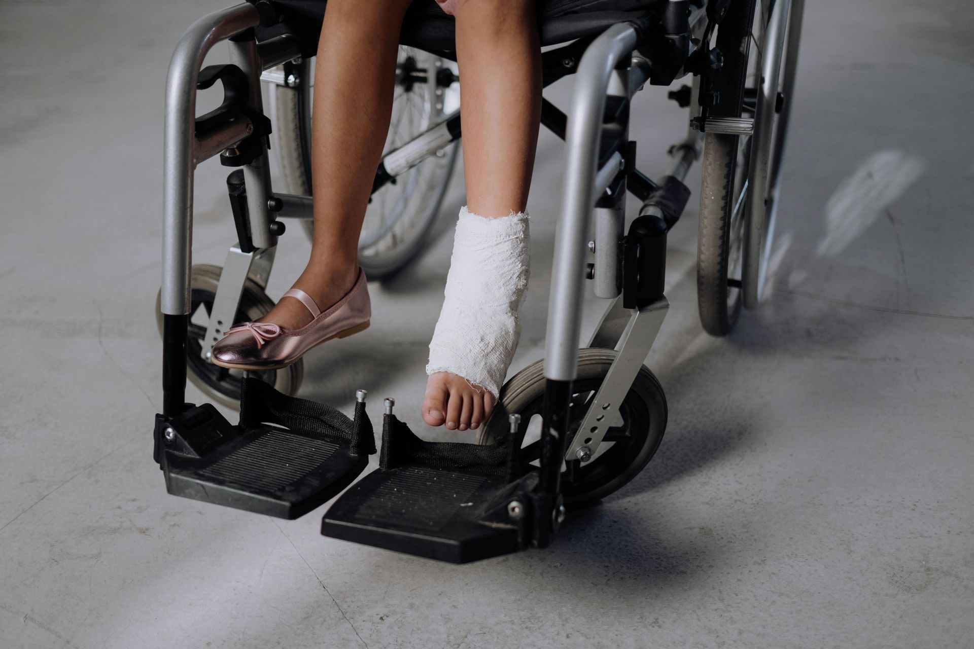 Person in a wheelchair with both feet in black leg braces, wearing a white cast on one ankle.