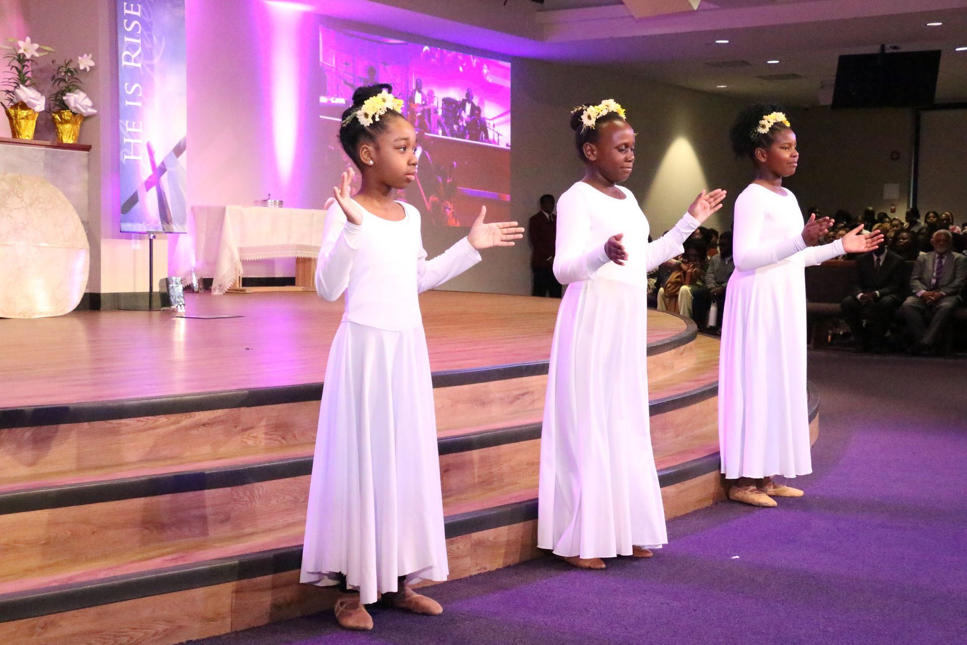 Three young girls in white dresses are standing on a stage.