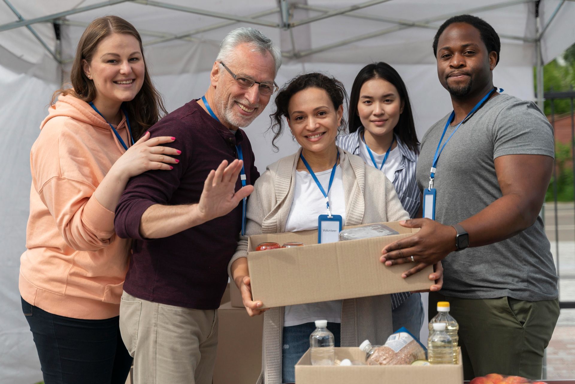 A diverse group of volunteers smiles while holding a cardboard box filled with food supplies under an outdoor tent.