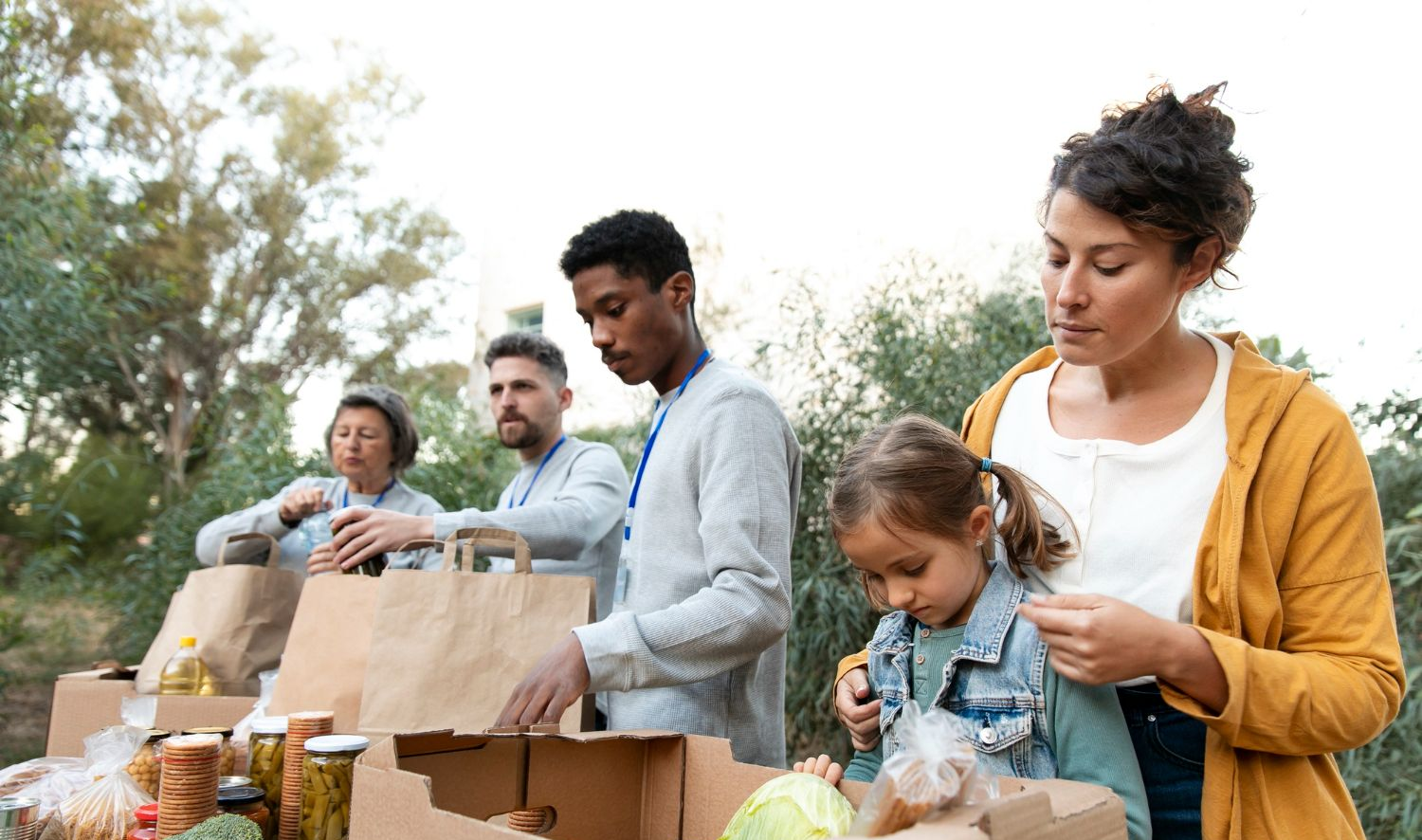 A diverse group of volunteers packs cardboard boxes with food items in an outdoor setting.