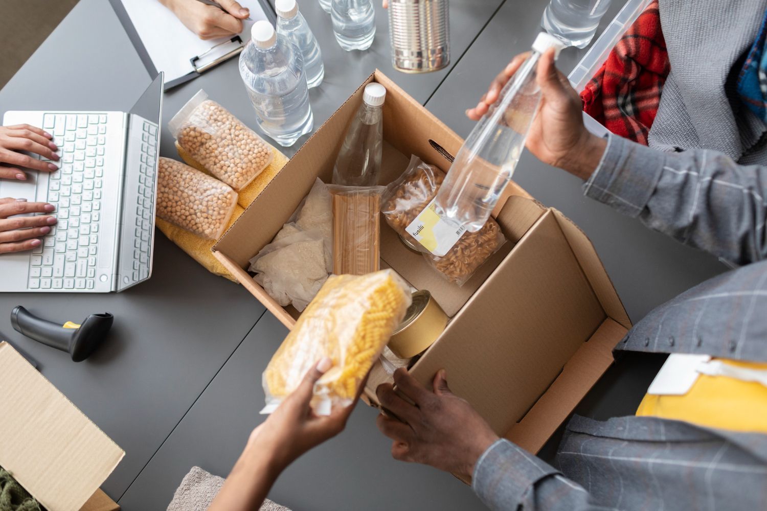 Volunteers packing food and water into a cardboard box on a table during a donation drive.