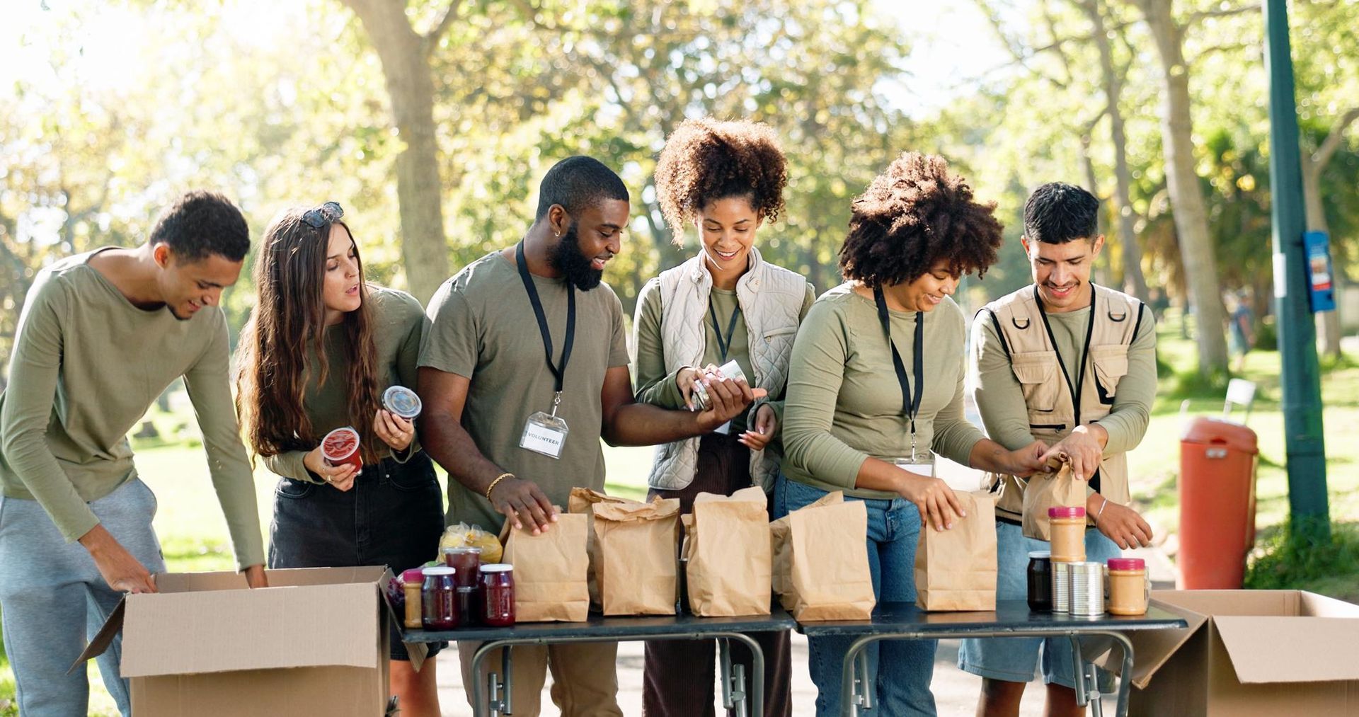 A diverse group of volunteers works outdoors at a park, filling brown paper bags with food at a community table.