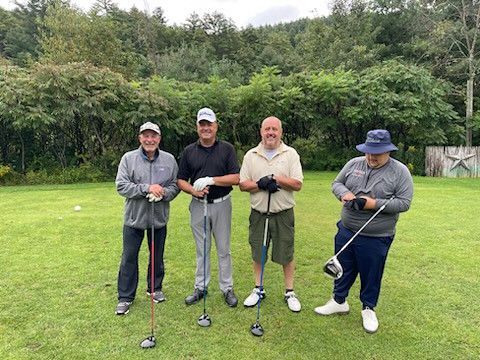 Four people pose on a grassy golf course, each holding a golf club, with a dense green treeline in the background.