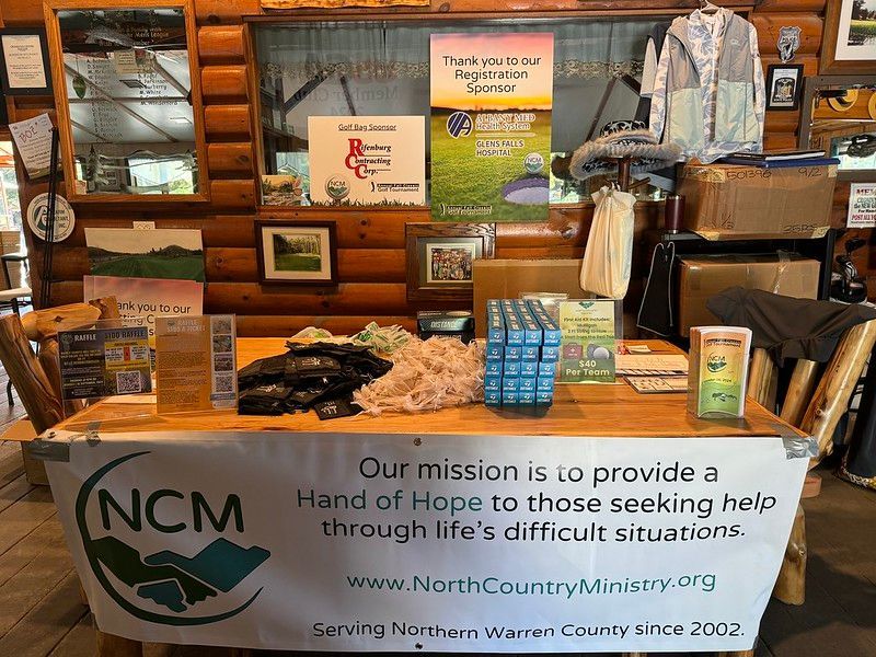 A table with a North Country Ministry banner, pamphlets, and supplies set up inside a rustic log cabin building.