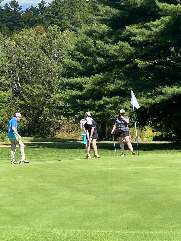 Three people on a golf green near the flag, preparing to putt on a sunny day with trees in the background.