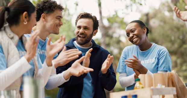 A diverse group of volunteers in matching blue shirts smile and clap outdoors at a community event.