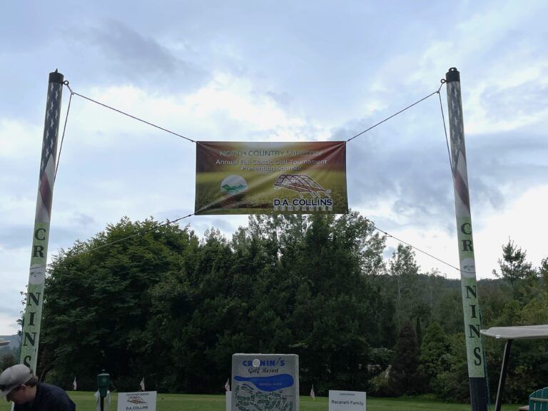 A banner suspended between two poles at a golf course, with a small course map on a sign in the foreground.