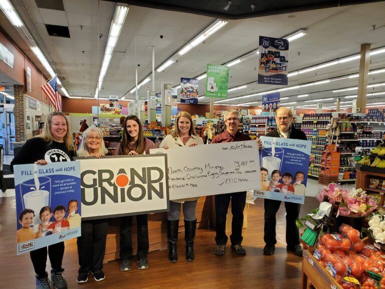 Six people stand in a Grand Union grocery store, holding a large donation check and promotional signs for a milk drive.