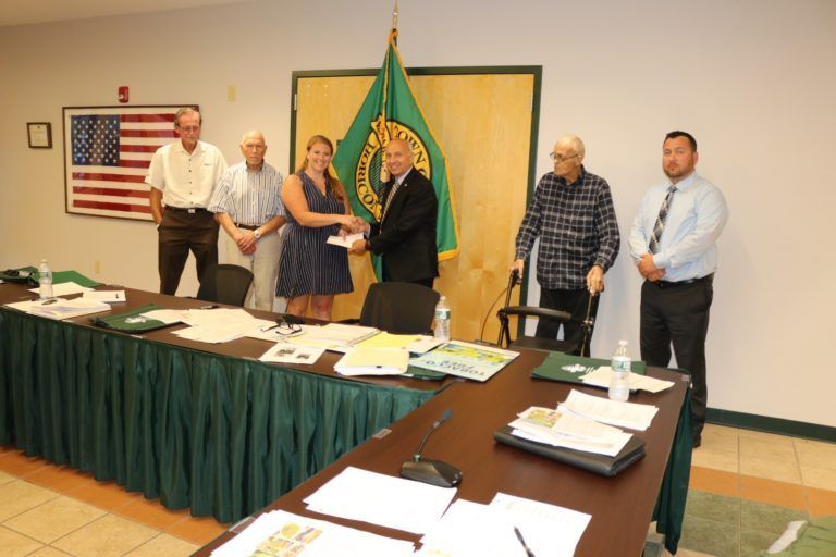 A woman in a blue dress shakes hands with a man in a suit in front of a flag, surrounded by observers in a meeting room.