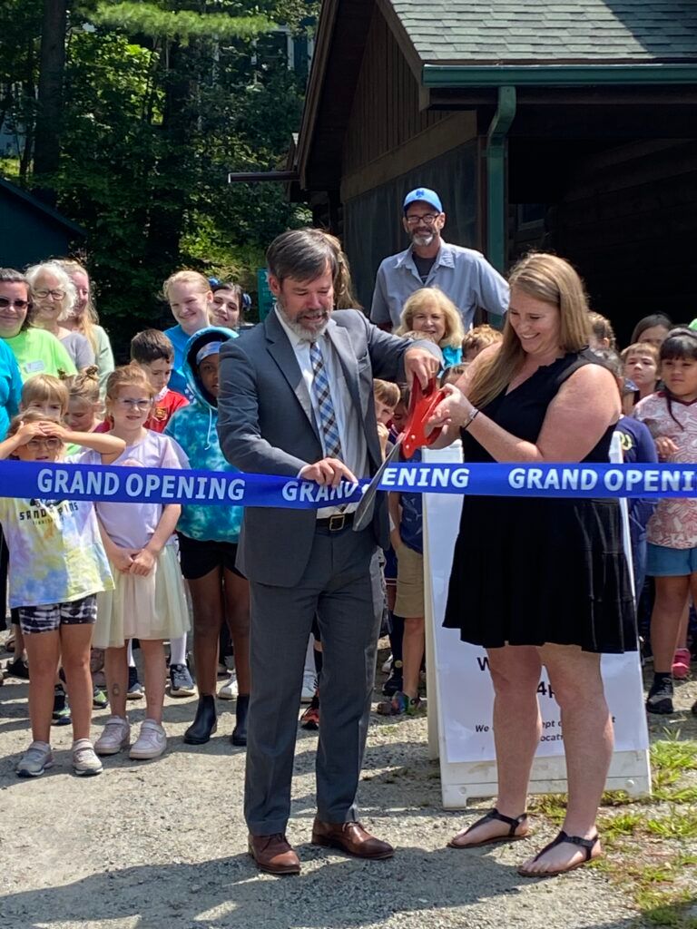 A man in a suit and a woman in a black dress cut a blue grand opening ribbon at an outdoor event surrounded by onlookers.