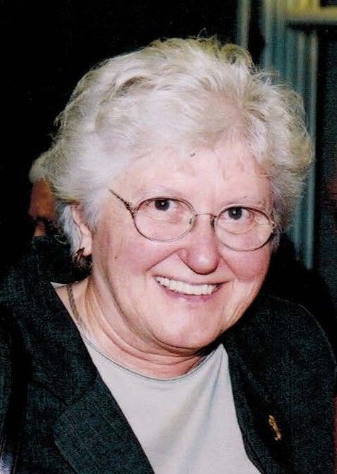 A smiling person with short white hair and glasses, wearing a dark jacket over a light-colored top.