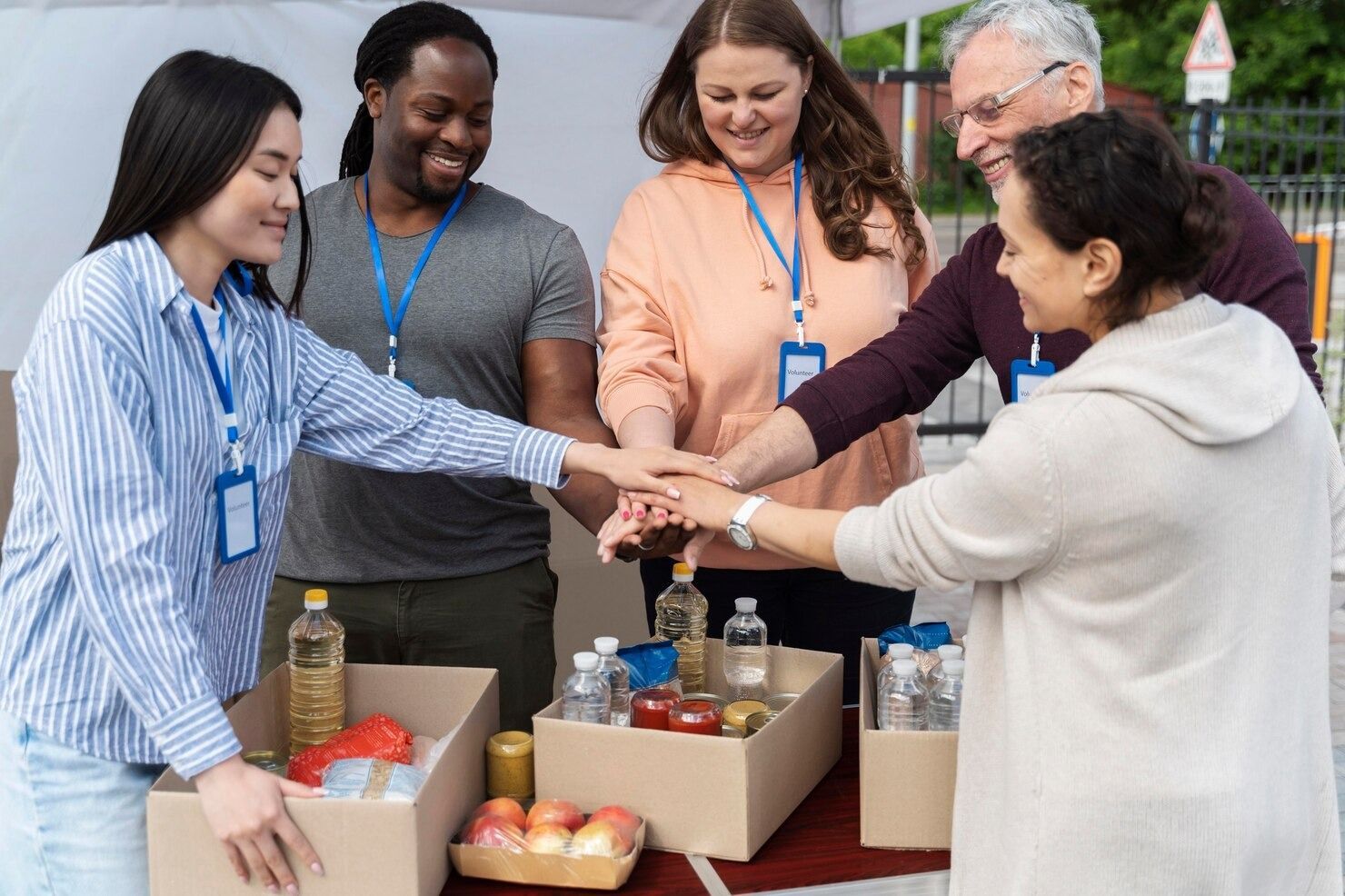 A group of diverse volunteers stacking their hands together over cardboard boxes filled with food items.