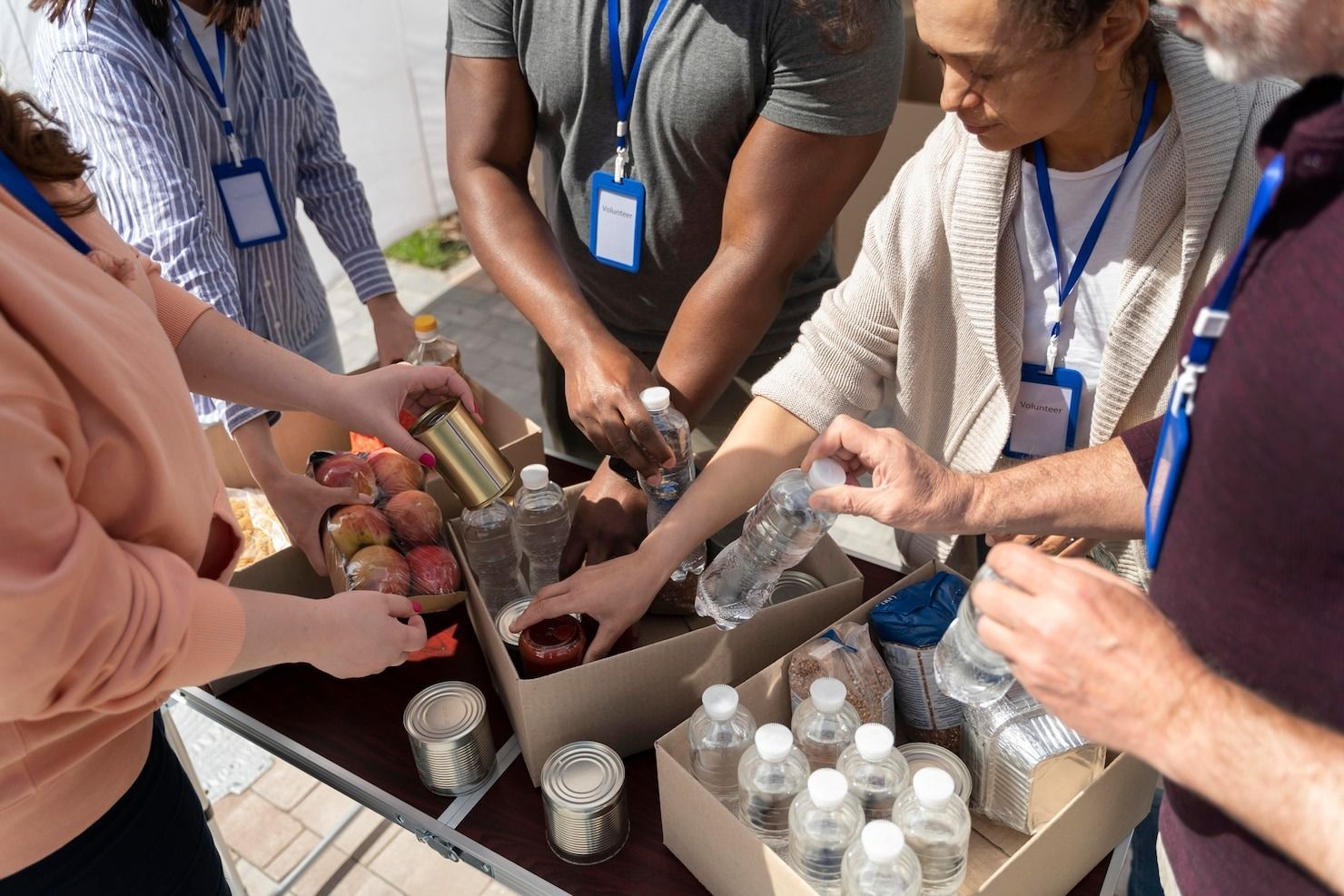 People with blue lanyards pack food items into cardboard boxes at a volunteer event.