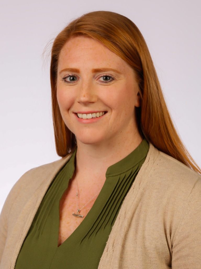 A smiling person with long reddish-brown hair, wearing an olive green top and a tan cardigan, against a light background.
