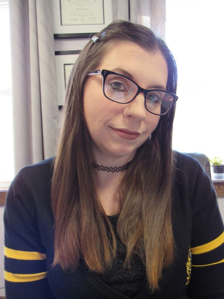 A person with long brown hair, wearing glasses and a black shirt with yellow stripes, looking toward the camera.
