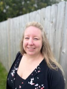 A person with long blonde hair smiles while standing outside in front of a wooden fence.