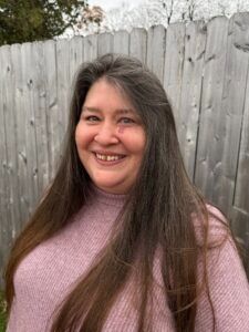 A person with long brown hair, wearing a pink sweater, smiles in front of a gray wooden fence outdoors.