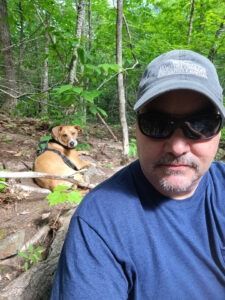 A person in a blue shirt, sunglasses, and baseball cap takes a selfie in a forest with a dog resting in the background.