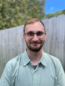A person with a beard and glasses smiles, wearing a light green collared shirt against a wooden fence backdrop.