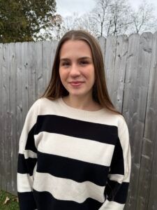 A person with long brown hair, wearing a black and white striped sweater, stands smiling in front of a wooden fence.