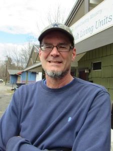 A person wearing a baseball cap and glasses smiling with arms crossed in front of a building with visible text.