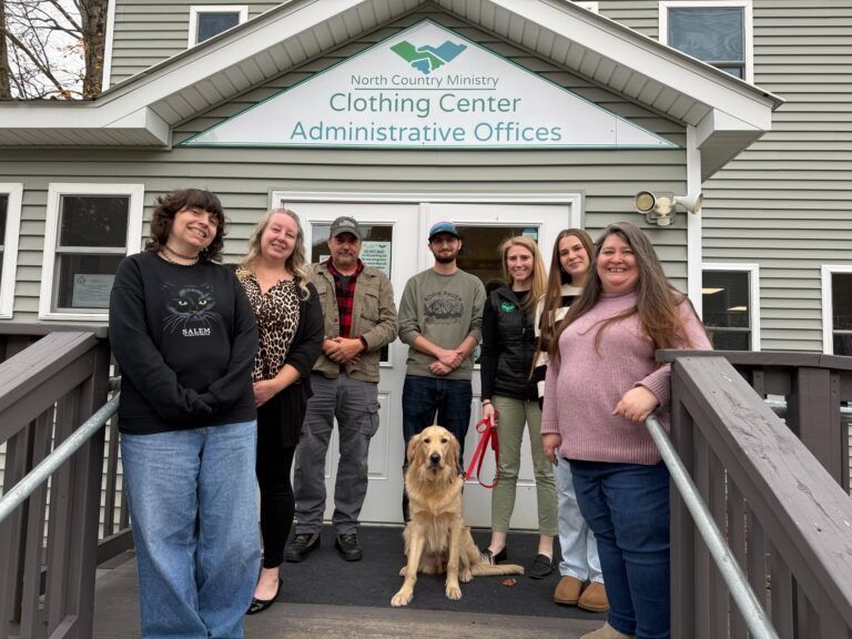 A group of eight people and a golden retriever stand smiling on the steps of the North Country Ministry building.