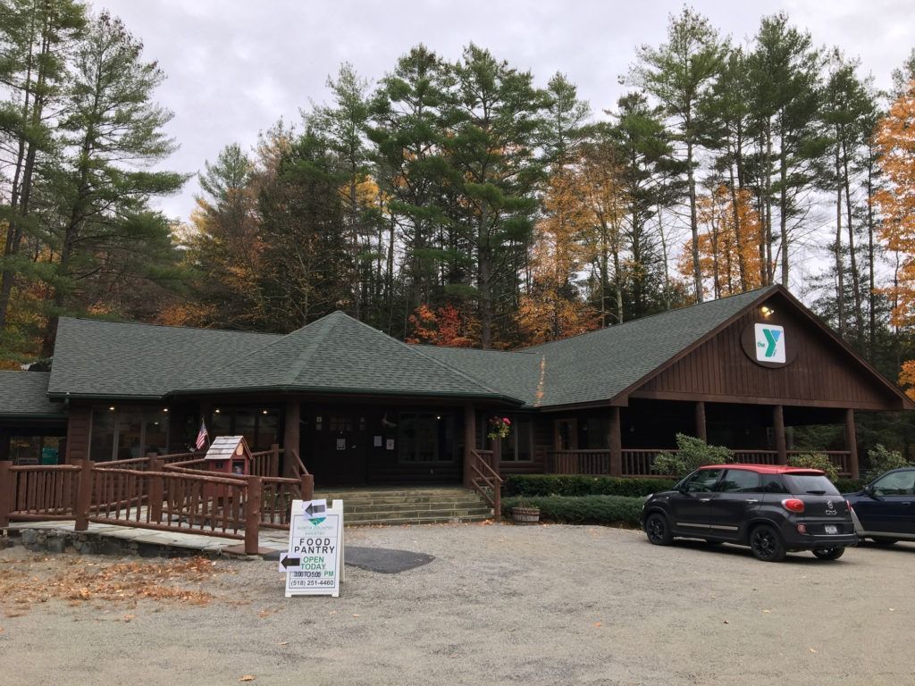 A rustic YMCA building with a green roof, wooden exterior, and a wheelchair ramp, set against an autumn forest.