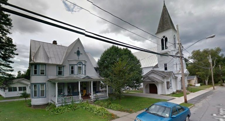 A white house with a wrap-around porch stands next to a white church with a steeple on a cloudy day.