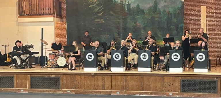A big band performs on a wooden stage in front of a painted forest backdrop, with musicians at stands and a drummer.