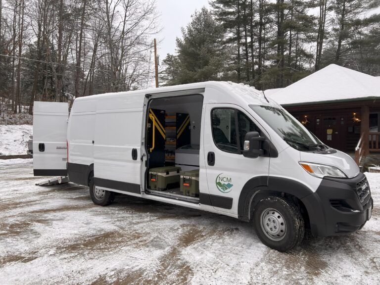 A white cargo van with its side and rear doors open, parked on a snow-covered lot near a building.