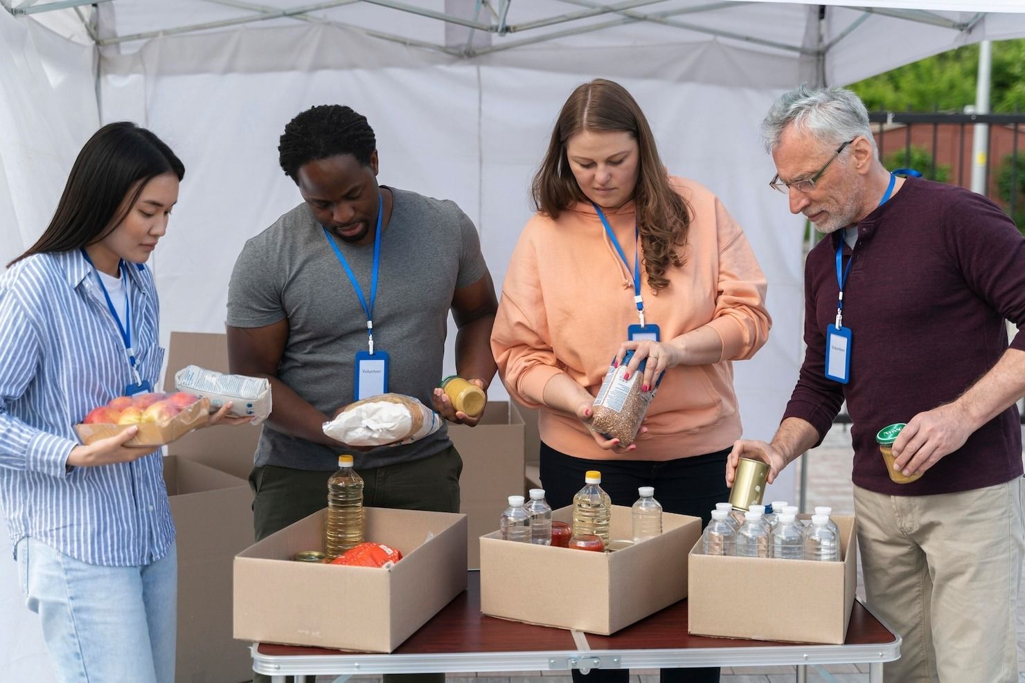 Four volunteers in casual clothing and lanyards sort food donations into cardboard boxes at a table under a white tent.