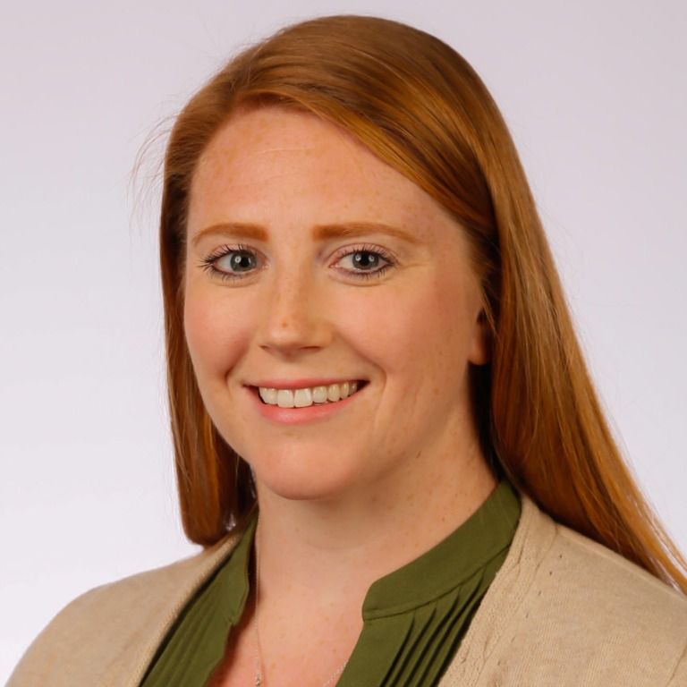 A professional portrait of a person with long red hair, smiling, wearing a green top and a beige cardigan.